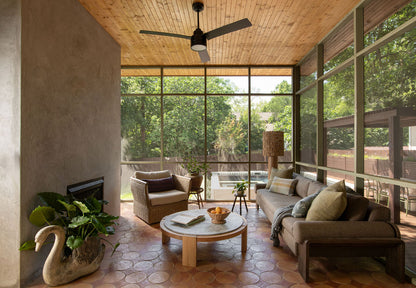 A spacious screened-in porch living room with a wood ceiling, large windows, terracotta tile flooring, a brown sofa with pillows, a matching armchair, a round coffee table, and a decorative swan-shaped planter with green plants.