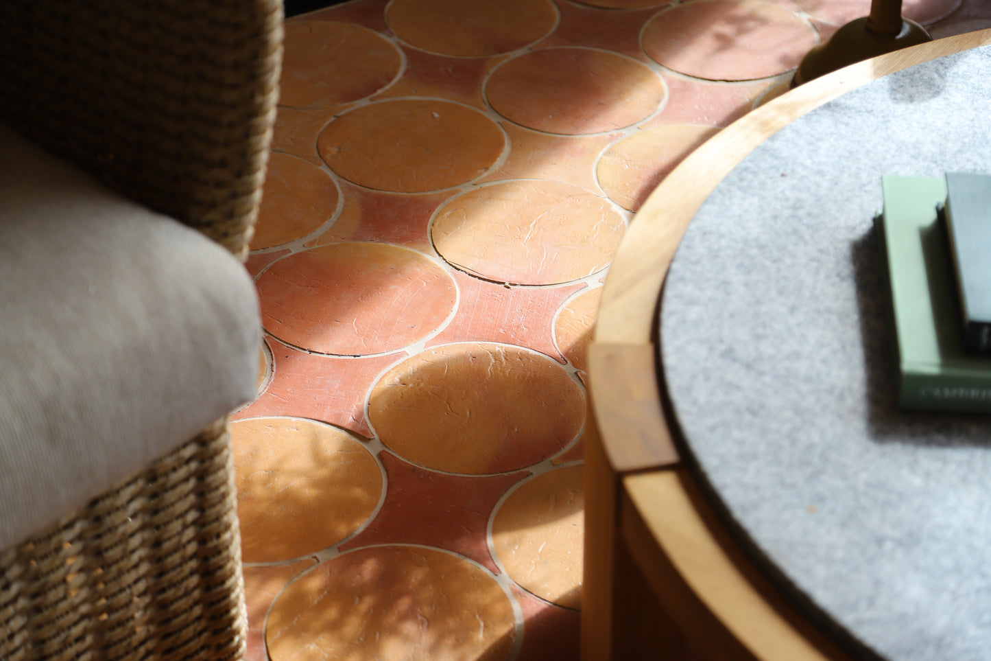 A close-up of terracotta-colored circular tile flooring with a wicker chair on the left and a round table on the right in a sunlit interior setting.