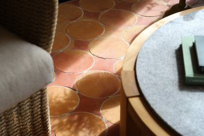 A close-up of terracotta-colored circular tile flooring with a wicker chair on the left and a round table on the right in a sunlit interior setting.