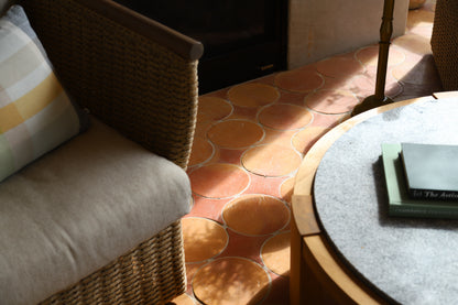 A close-up lifestyle shot of a wicker armchair with a beige cushion and a striped pillow, next to a round gray-top table on terracotta tile flooring.