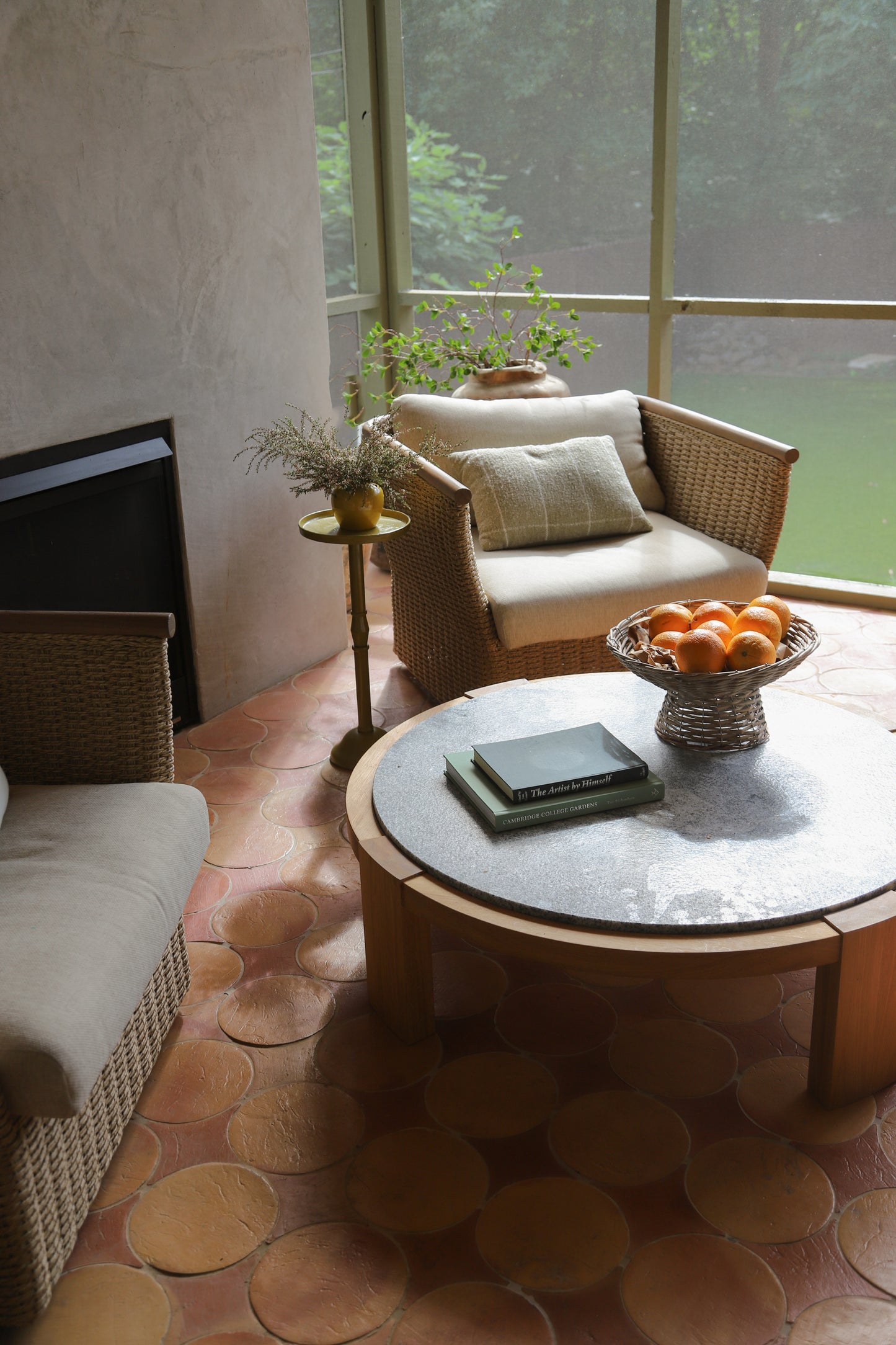 A cozy indoor patio or screened room corner with woven wicker furniture, a round stone-top coffee table, a bowl of oranges, and a large window looking out onto greenery.