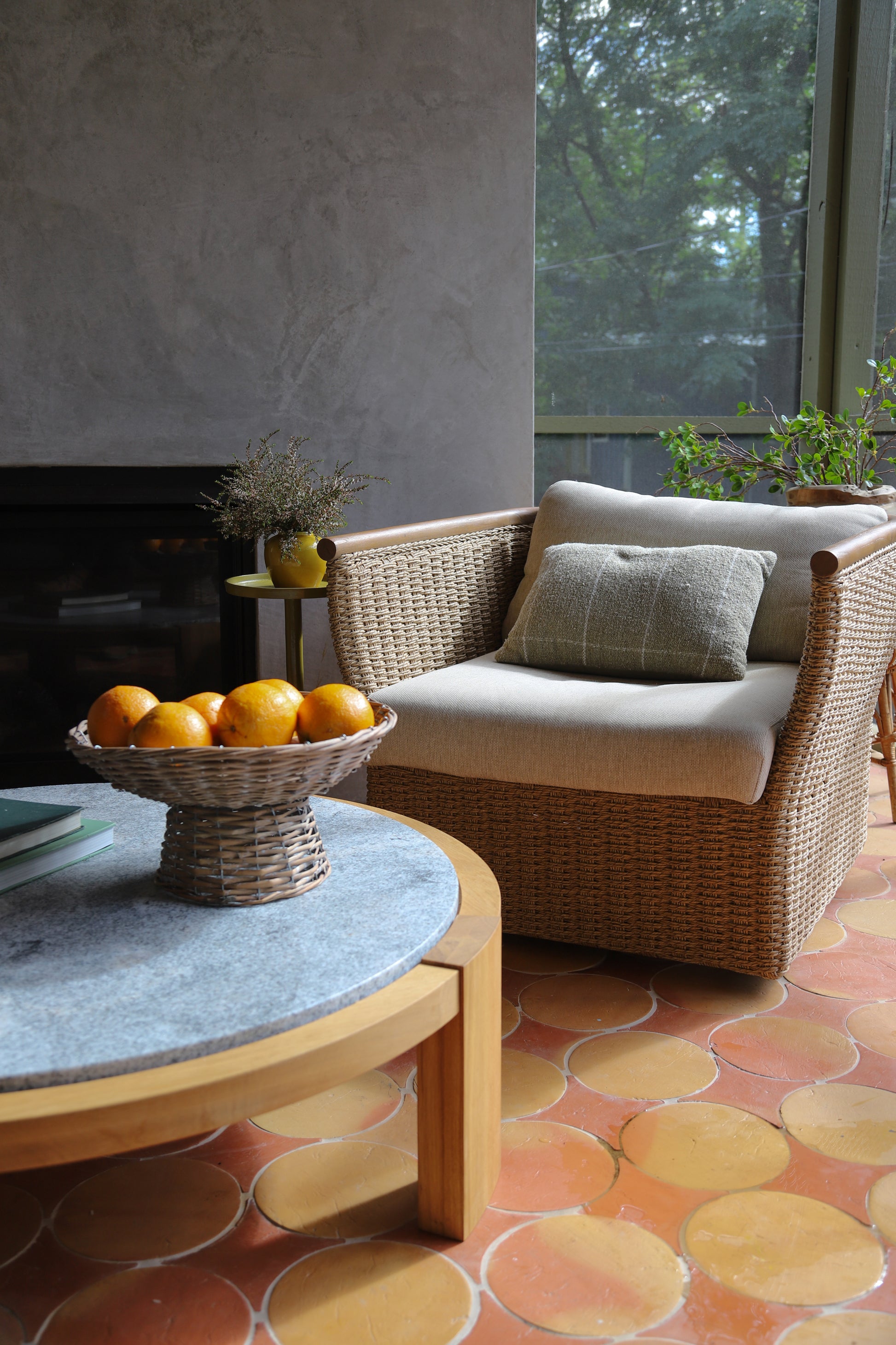 A woven wicker armchair with cream cushions and a matching throw pillow, placed in a rustic living room setting with a stone-topped coffee table, oranges, and a terracotta patterned tile floor.