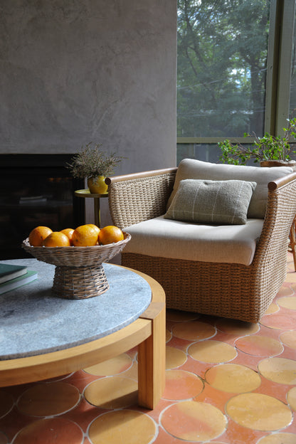 A woven wicker armchair with cream cushions and a matching throw pillow, placed in a rustic living room setting with a stone-topped coffee table, oranges, and a terracotta patterned tile floor.