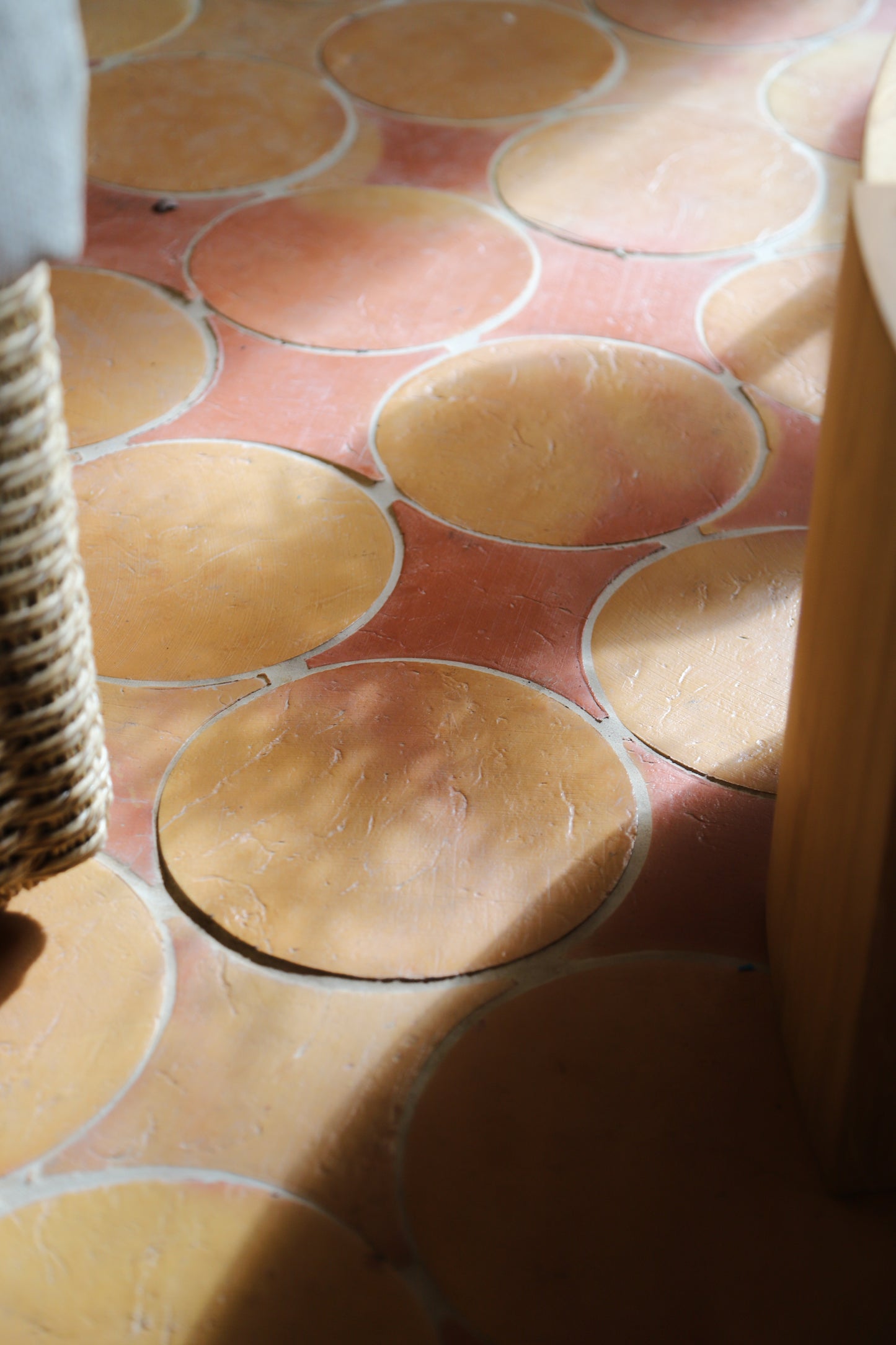 Close-up of terracotta-colored circular floor tiles with a warm pinkish-red grout or border, partially framed by a woven wicker object on the left and a wooden structure on the right.