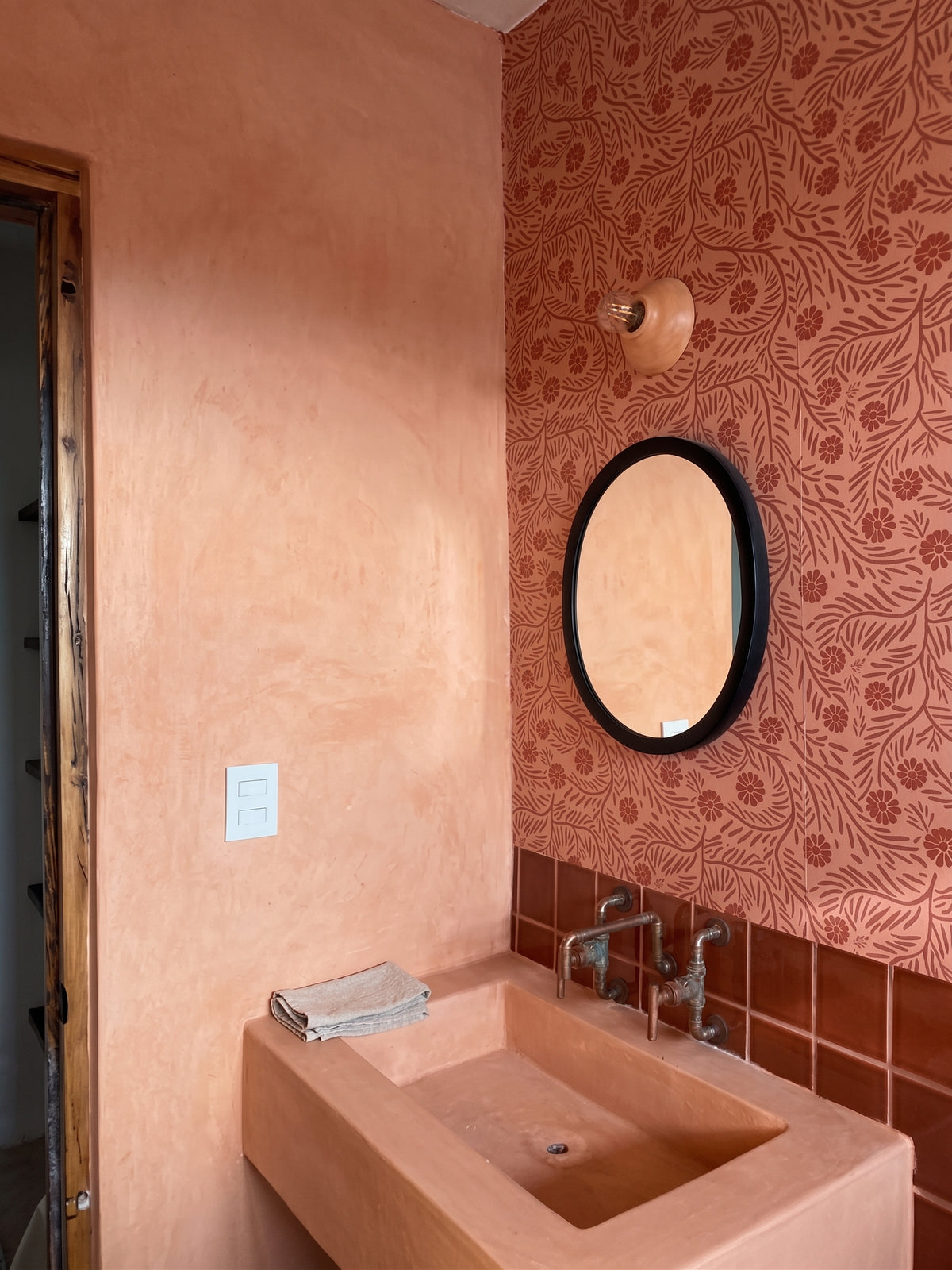 A terracotta-colored wall-mounted sink with a matching rectangular basin, set against a textured terracotta plaster wall and a patterned wallpapered accent wall with an oval mirror.