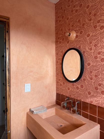 A terracotta-colored wall-mounted sink with a matching rectangular basin, set against a textured terracotta plaster wall and a patterned wallpapered accent wall with an oval mirror.