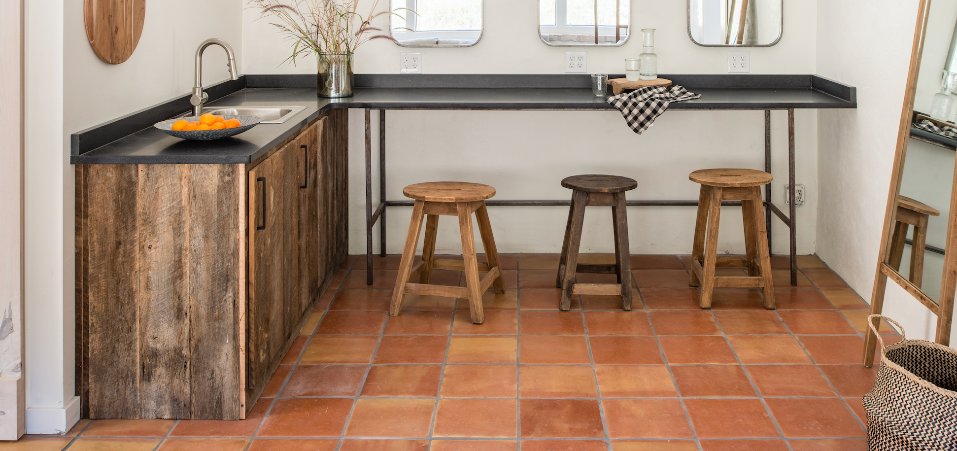 Rustic L-shaped kitchen island with weathered wood cabinets, a black countertop, a sink, and three wooden stools on terracotta tile flooring.