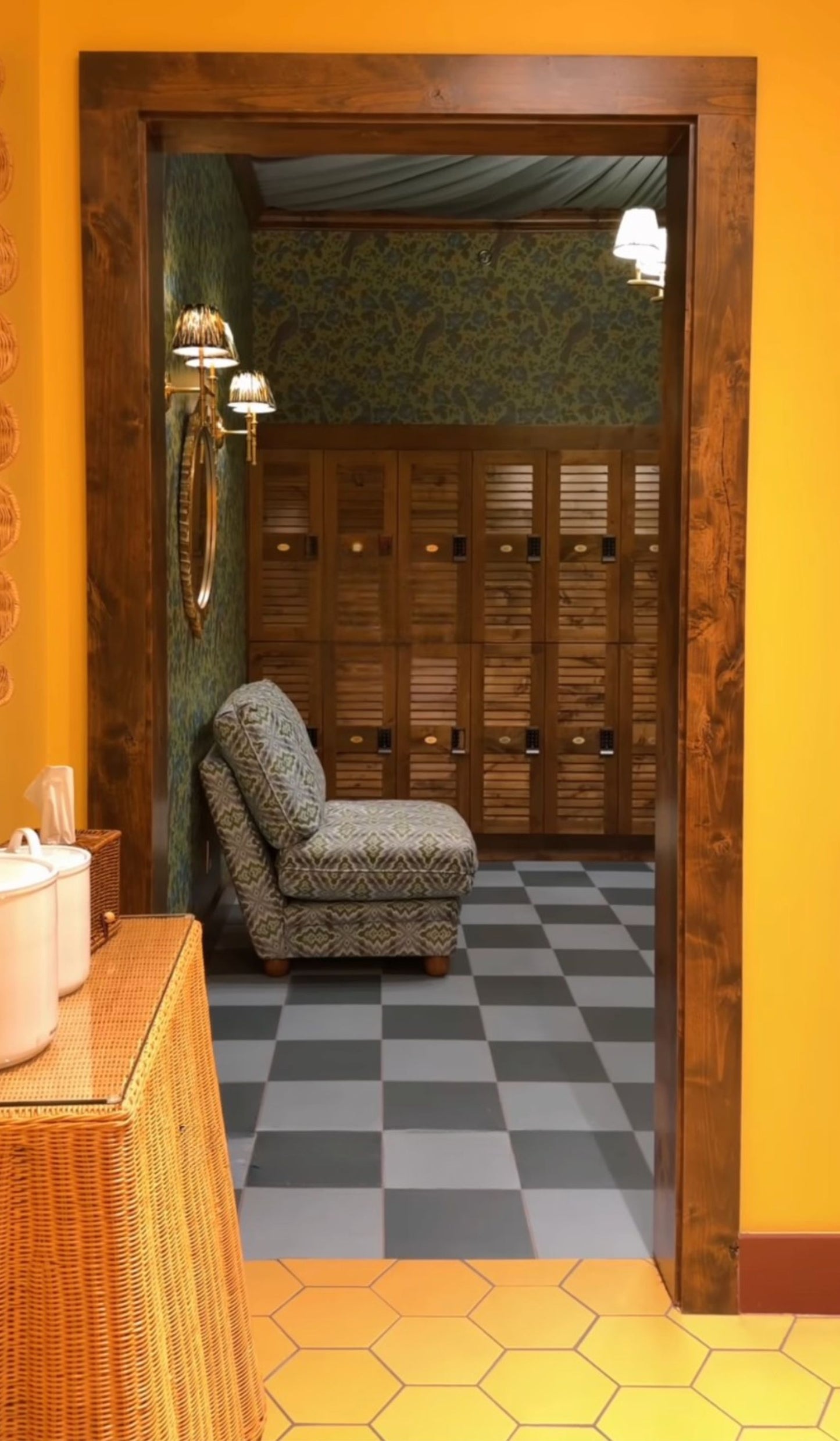 A warm interior doorway with a dark wood frame, bright yellow walls, and a checkered gray and white tile floor. Through the opening, a patterned armchair sits in front of wooden locker-style cabinets, with a wicker side table and white containers visible in the foreground.