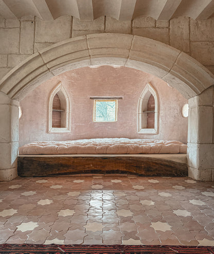 A rustic stone room with a built-in cushioned daybed under a large arched opening, featuring terracotta tiled floors and a pink plaster wall.