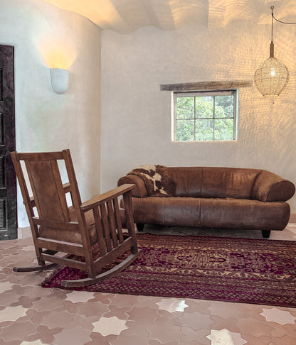 A rustic brown leather sofa and a dark wood rocking chair in a styled room with textured plaster walls, a patterned rug, and star-shaped floor tiles.