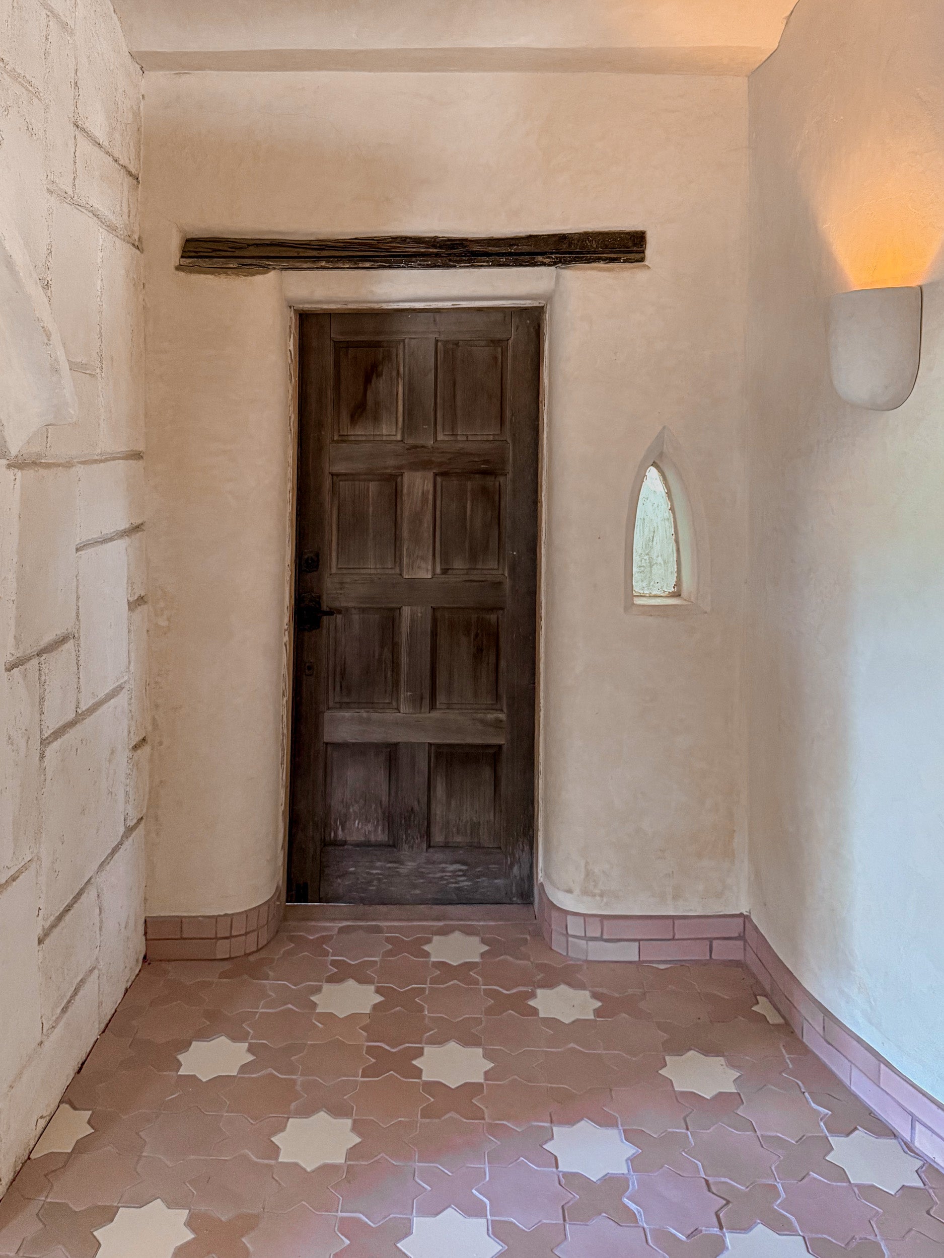 A rustic interior hallway with a dark wooden door centered on a beige plaster wall, patterned terracotta and cream floor tiles, a small arched window, and a wall-mounted light.