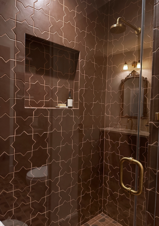 A luxury walk-in shower featuring dark brown mosaic tiles with a raised geometric gold pattern, a built-in niche with a soap dish, and a gold-framed glass door with a round pull handle.