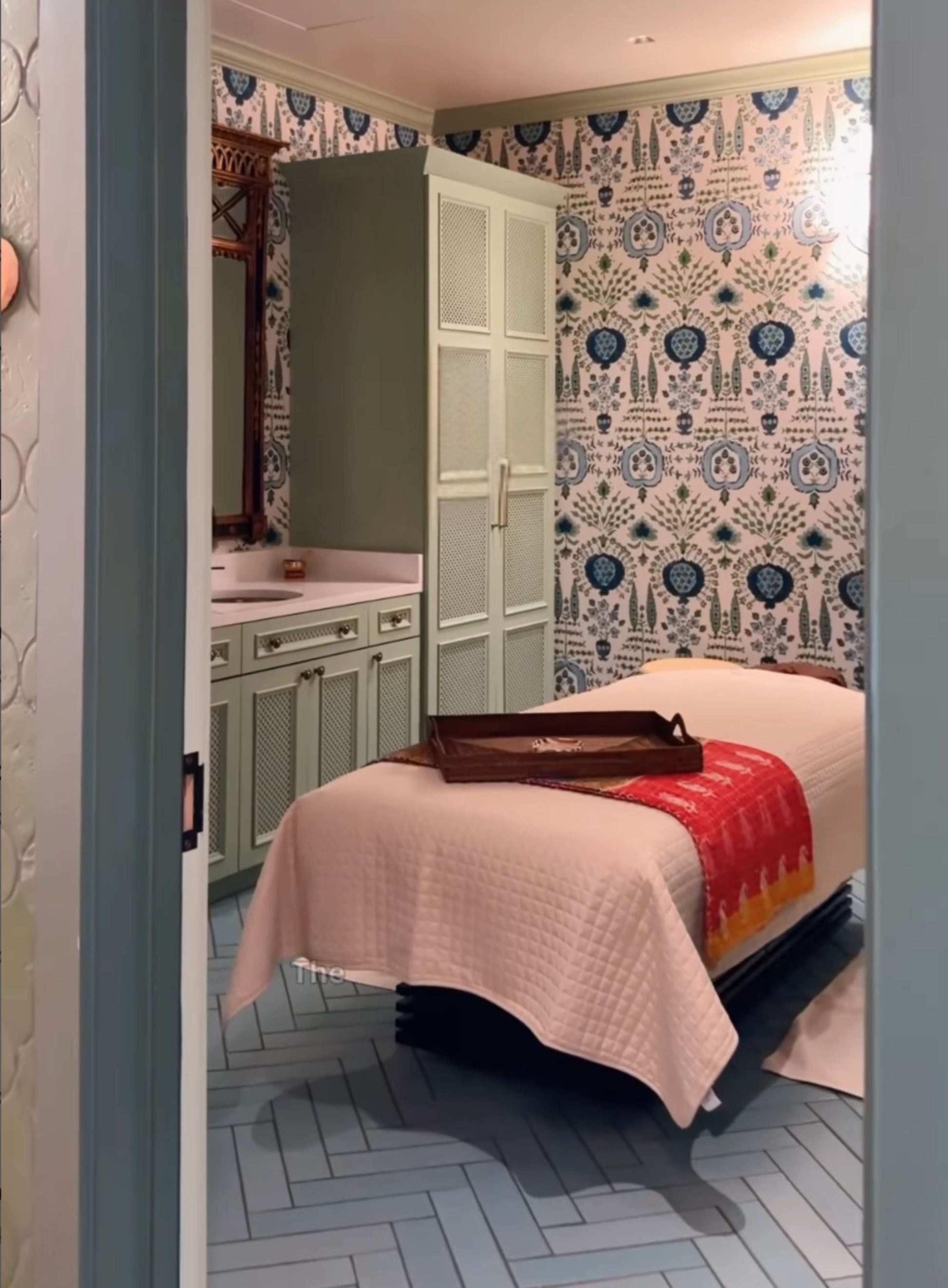 A spa treatment room with a massage table covered in a cream quilted blanket and a red-orange throw, set against patterned blue and white wallpaper and light green cabinetry.
