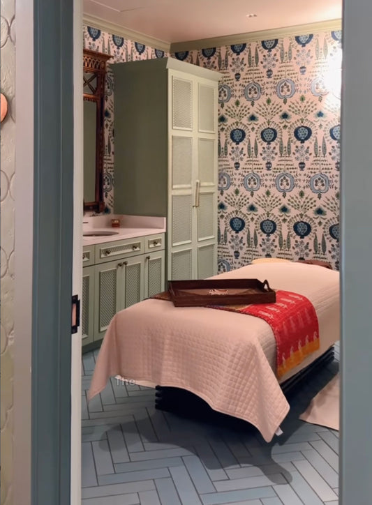 A spa treatment room with a massage table covered in a cream quilted blanket and a red-orange throw, set against patterned blue and white wallpaper and light green cabinetry.