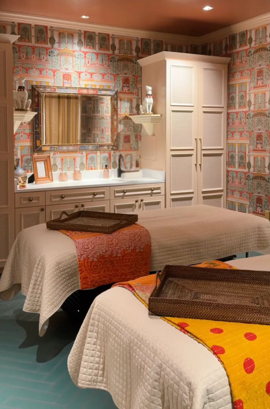 A luxurious spa or massage room featuring two treatment beds with cream and yellow patterned quilts, a vanity with a mirror, and a tall white cabinet, all set against ornate multicolor patterned wallpaper.