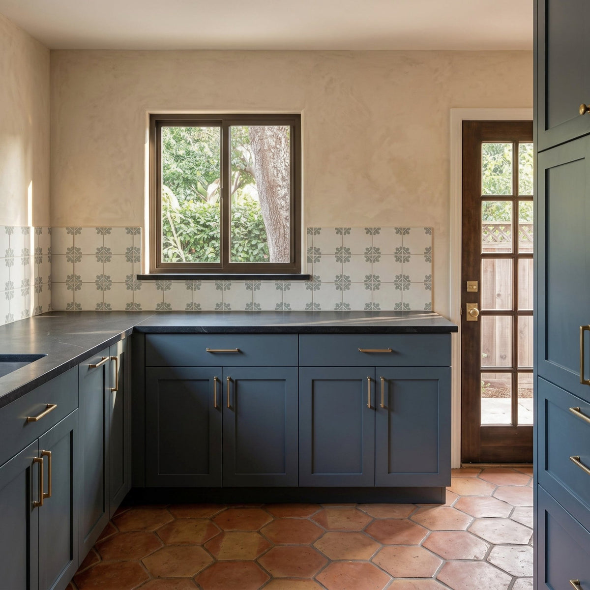 A kitchen interior with dark blue cabinets, a black countertop, patterned tile backsplash, terracotta hexagonal floor tiles, a window, and a glass-paneled door.