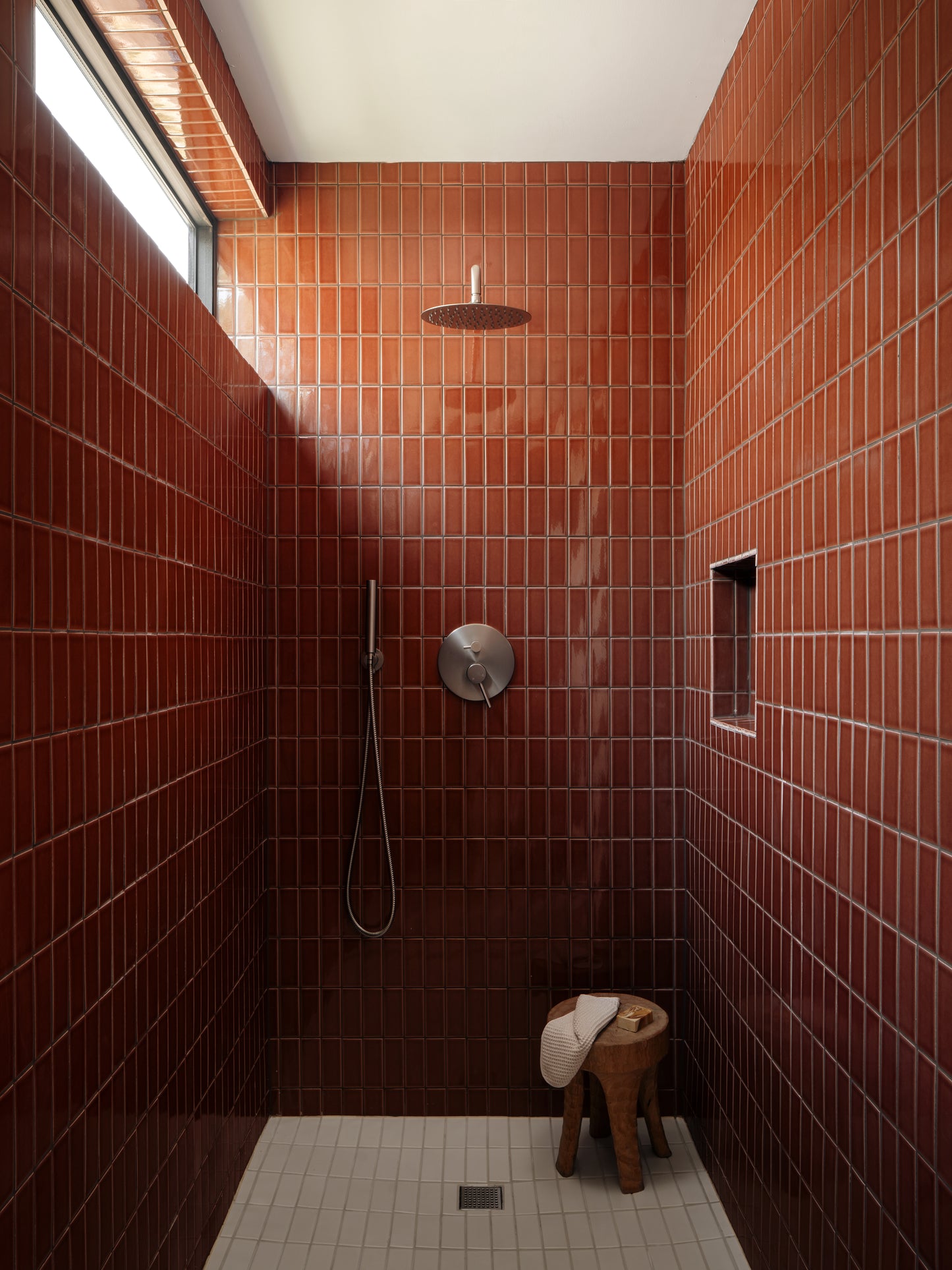 A minimalist walk-in shower stall with glossy reddish-brown rectangular tiles, a round silver shower head, a handheld shower hose, a small wooden stool with a white towel, and a tiled floor.