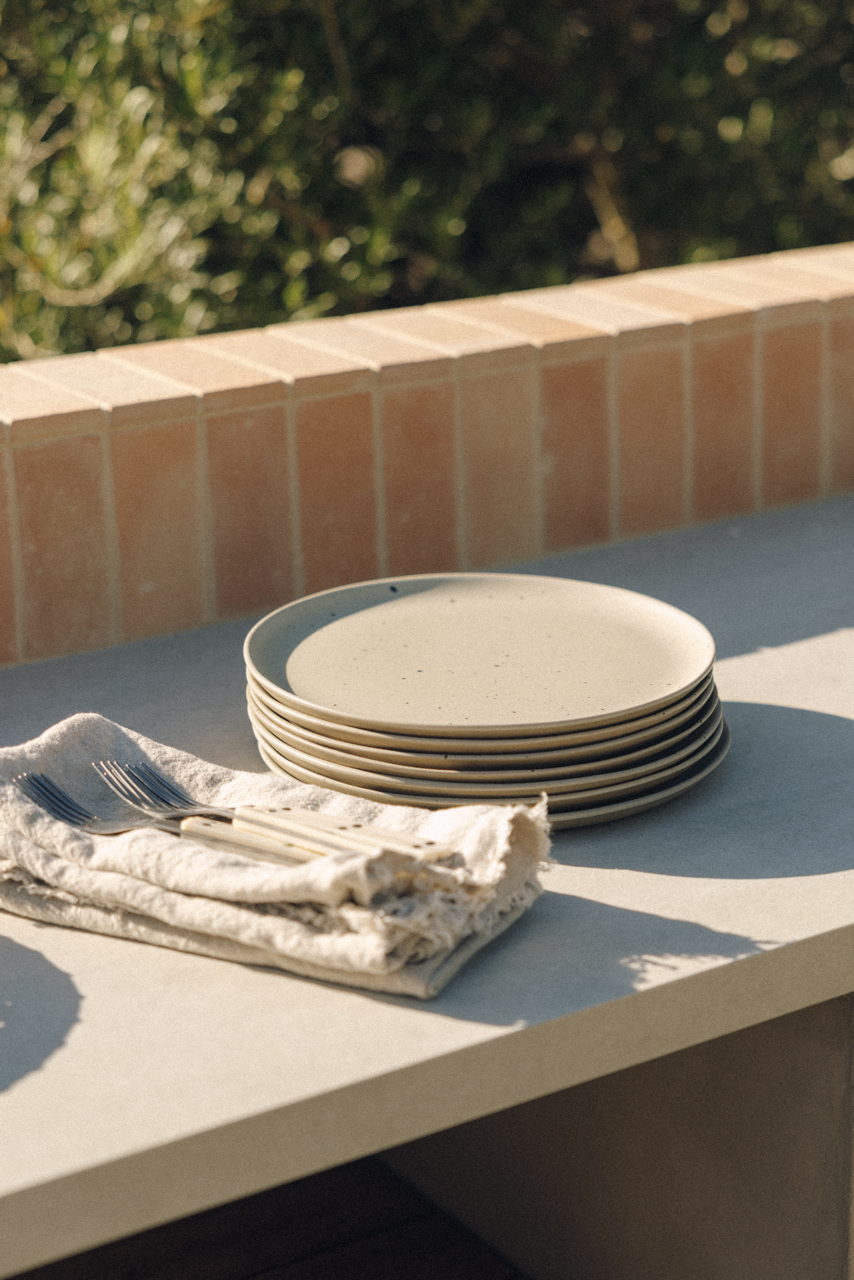 A stack of speckled beige ceramic plates sits on a light stone table next to a folded cream cloth napkin with silver cutlery, with a terracotta tile wall and blurred green foliage in the background.