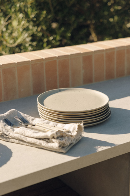 A stack of speckled beige ceramic plates sits on a light stone table next to a folded cream cloth napkin with silver cutlery, with a terracotta tile wall and blurred green foliage in the background.