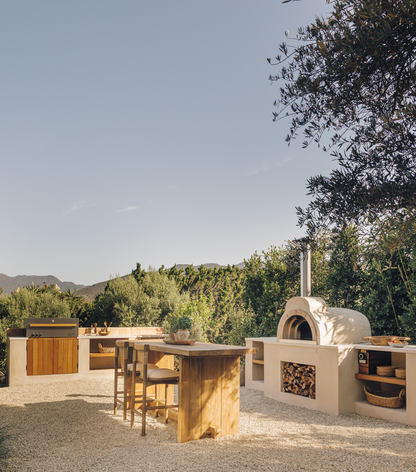 Outdoor kitchen setup with a white stone pizza oven, built-in grills, a central wooden island, and bar stools, set on a gravel surface with trees and hills in the background.