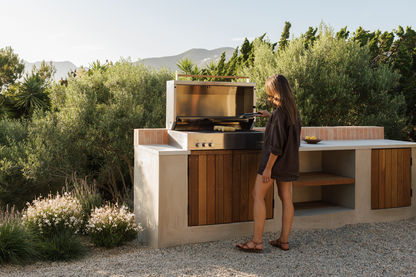 A woman stands beside an outdoor built-in kitchen island with a stainless steel gas grill, set against a backdrop of green shrubs and distant mountains.