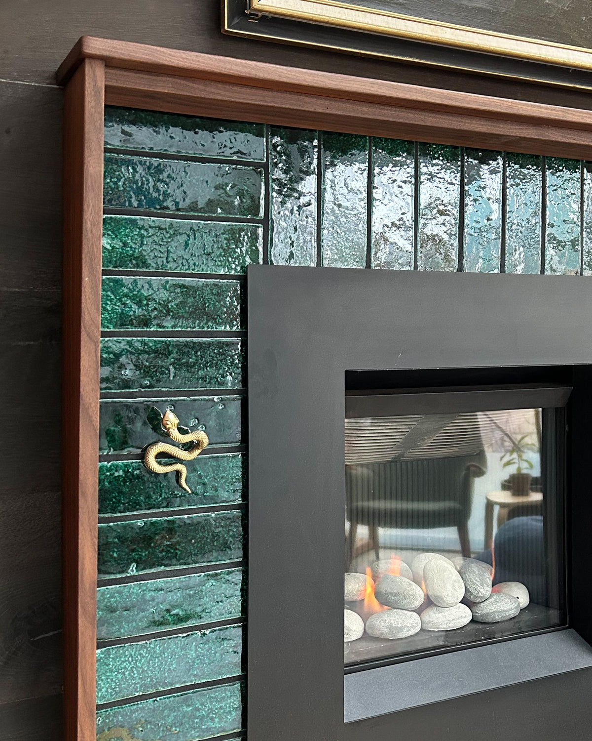 A small gold snake figurine clings to the glossy emerald green glazed ceramic tile surrounding a modern fireplace with a gas flame and grey stone pebbles.