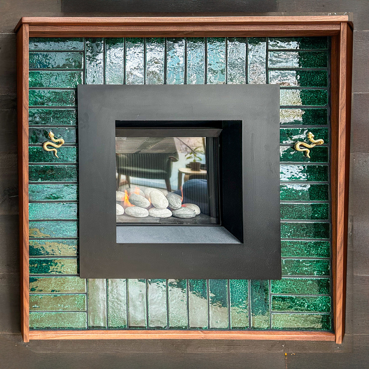 A modern gas fireplace features a square black metal surround framed by glossy green ceramic tiles and two decorative brass snakes.
