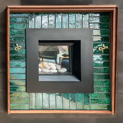 A modern gas fireplace features a square black metal surround framed by glossy green ceramic tiles and two decorative brass snakes.