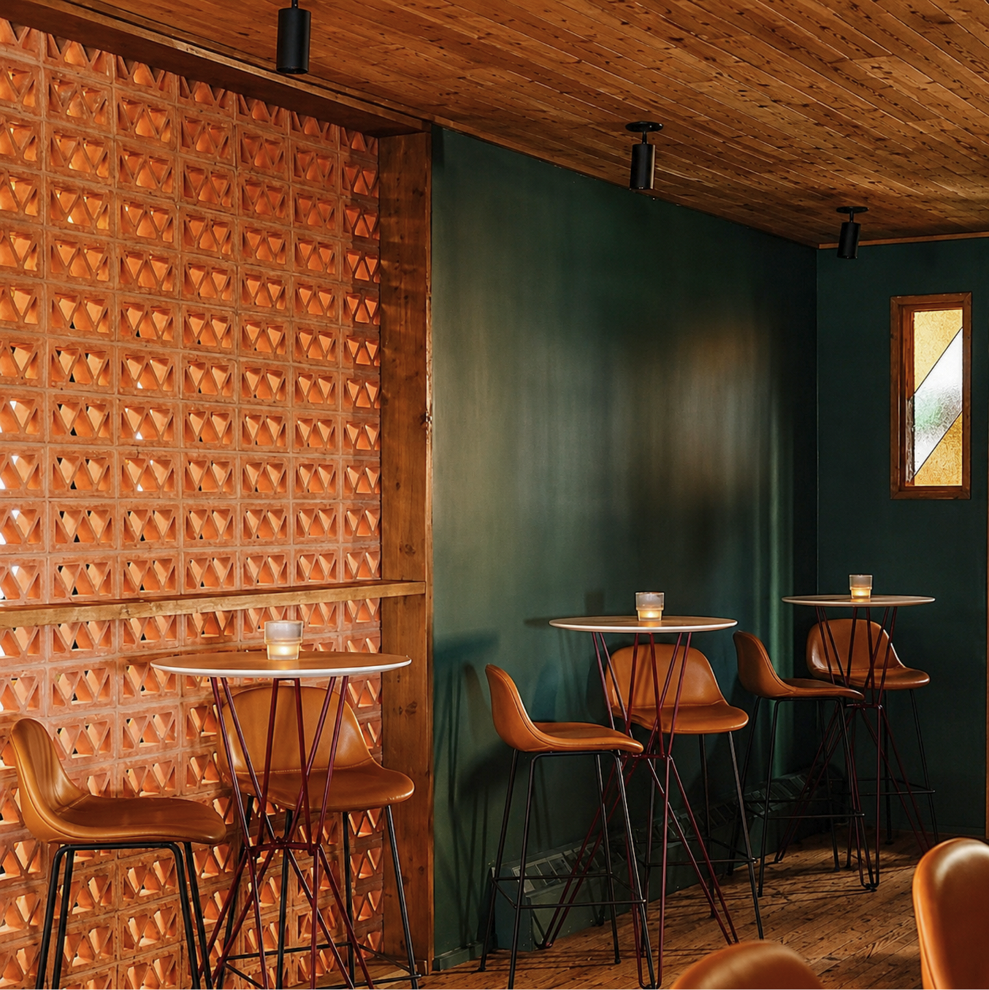 A modern interior scene with tall bar-height tables and brown leather stools in a restaurant or cafe setting. A textured terracotta tile wall is prominent on the left, with dark green and wood-paneled walls and ceiling visible.