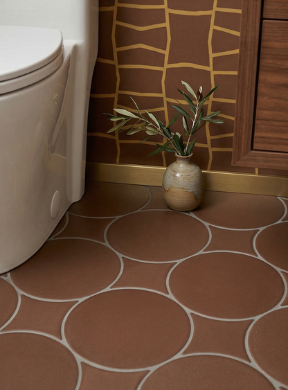 Terracotta brown solid circular tiles by Clay Imports, with white grout, laid on a bathroom floor next to a toilet and a vase of olive branches.