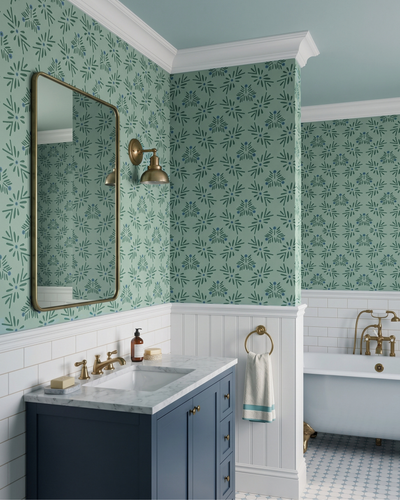A styled bathroom interior featuring green and white patterned wallpaper, white wainscoting, a blue vanity, a large gold-framed mirror, brass fixtures, and a white clawfoot bathtub in the background.