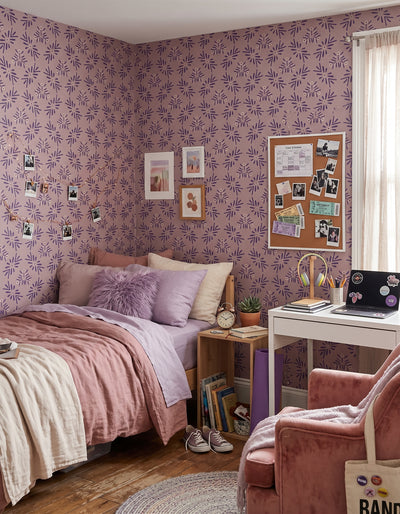 A cozy bedroom corner with lavender and purple floral wallpaper, a bed with pink and purple bedding, a white desk with a laptop, a pink armchair, and a cork bulletin board on the wall.