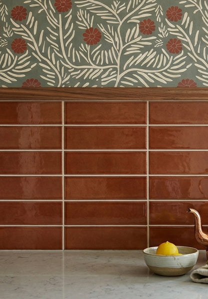 Glossy reddish-brown elongated solid tiles by Clay Imports installed as a kitchen backsplash, with botanical wallpaper above and lemons on a countertop.