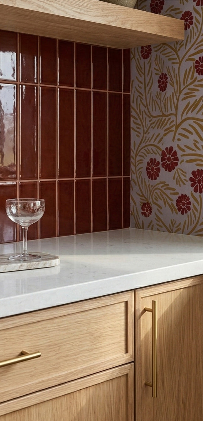 Burgundy gloss 2x8 solid tile by Clay Imports, vertically installed as a kitchen backsplash next to floral wallpaper and above wood cabinets.
