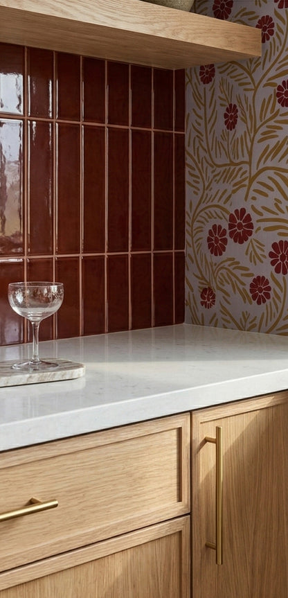 Burgundy gloss 2x8 solid tile by Clay Imports, vertically installed as a kitchen backsplash next to floral wallpaper and above wood cabinets.