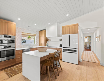 A modern kitchen interior with light wood cabinets, a white island, stainless steel appliances, a white refrigerator, and wood-look bar stools, shown in a real home environment.