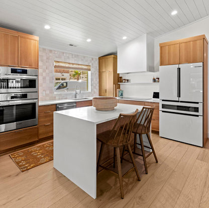 A modern kitchen interior with a white island, wood bar stools, light wood flooring, white shiplap ceiling, stainless steel appliances, and light wood cabinetry.
