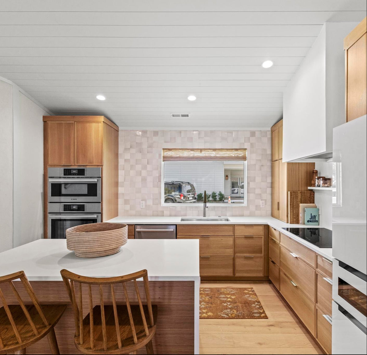 A modern kitchen interior with light wood cabinetry, a white island, two wooden chairs, built-in stainless steel appliances, and a white refrigerator.
