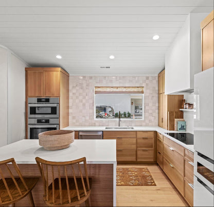 A modern kitchen interior with light wood cabinetry, a white island, two wooden chairs, built-in stainless steel appliances, and a white refrigerator.