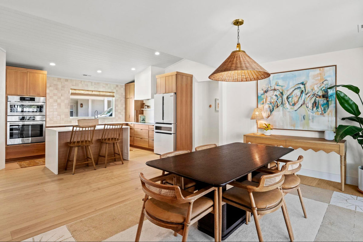 A modern open-plan dining and kitchen interior with a dark wood dining table, six woven-back chairs, a wicker pendant light, light oak cabinetry, a tiled island backsplash, and a large abstract painting on the wall.