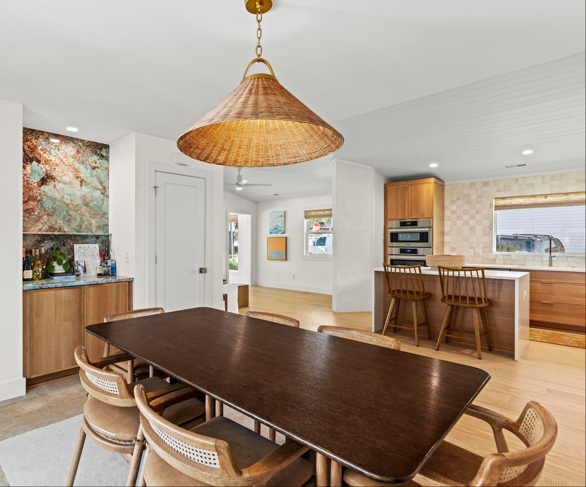 A dining room scene with a dark wood rectangular table and six woven-back chairs under a large conical woven pendant light. The room has light wood flooring, white walls, an open kitchen area with wood cabinetry and bar stools, and a turquoise accent wall in the adjacent room.