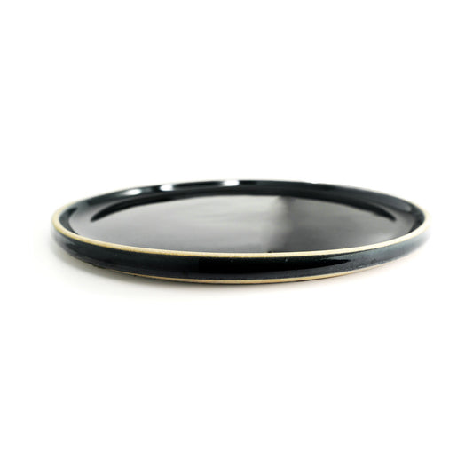 Black dinner plate by Clay Imports with a glossy finish and tan rim, viewed at a low angle.
